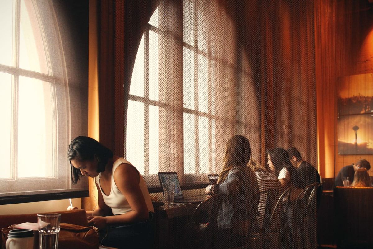 A woman working on a laptop in a warmly lit cafe beside a tall arched window