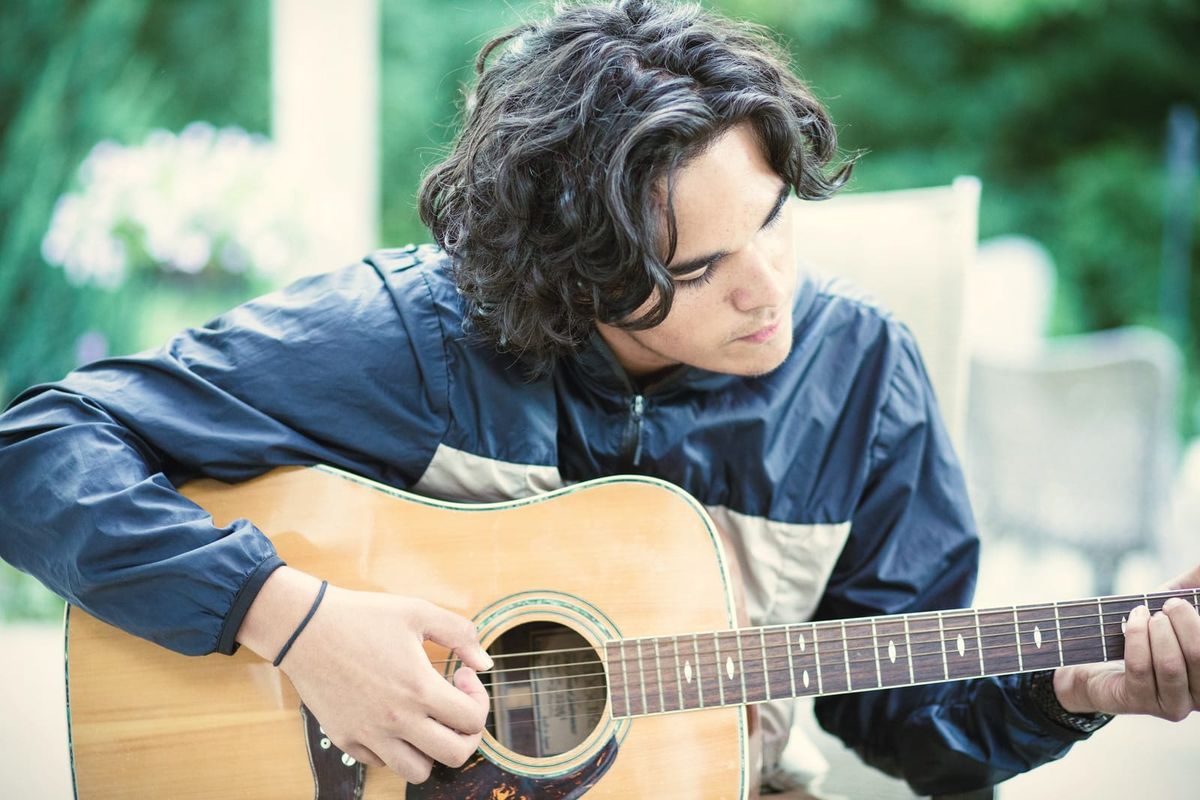 A young man playing an acoustic guitar outdoors, eyes on the strings