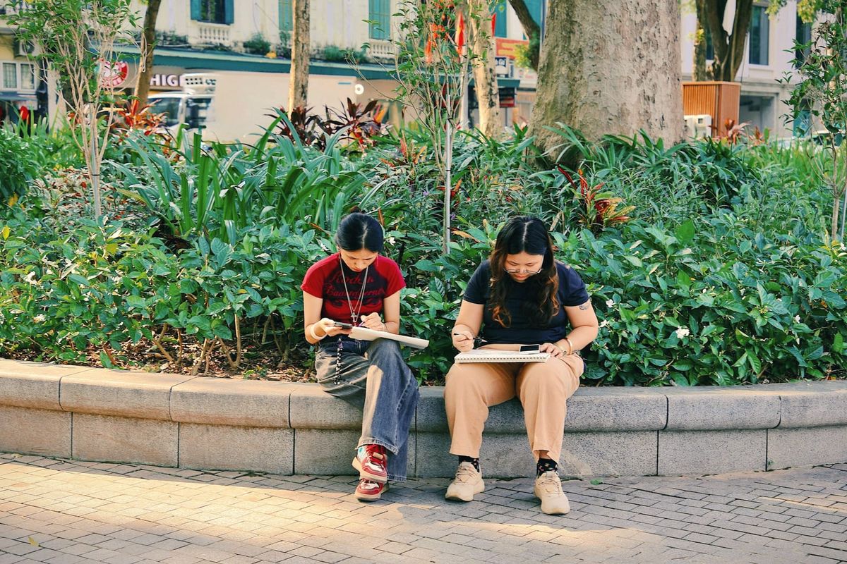 Two young women sketching on a low wall among green plants in a park