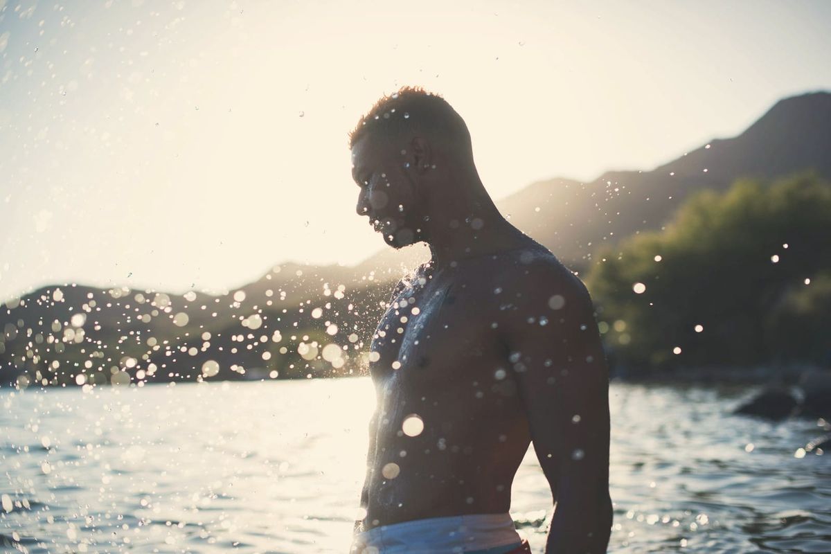 A man in profile mid-splash beside a mountain lake, backlit by the sun