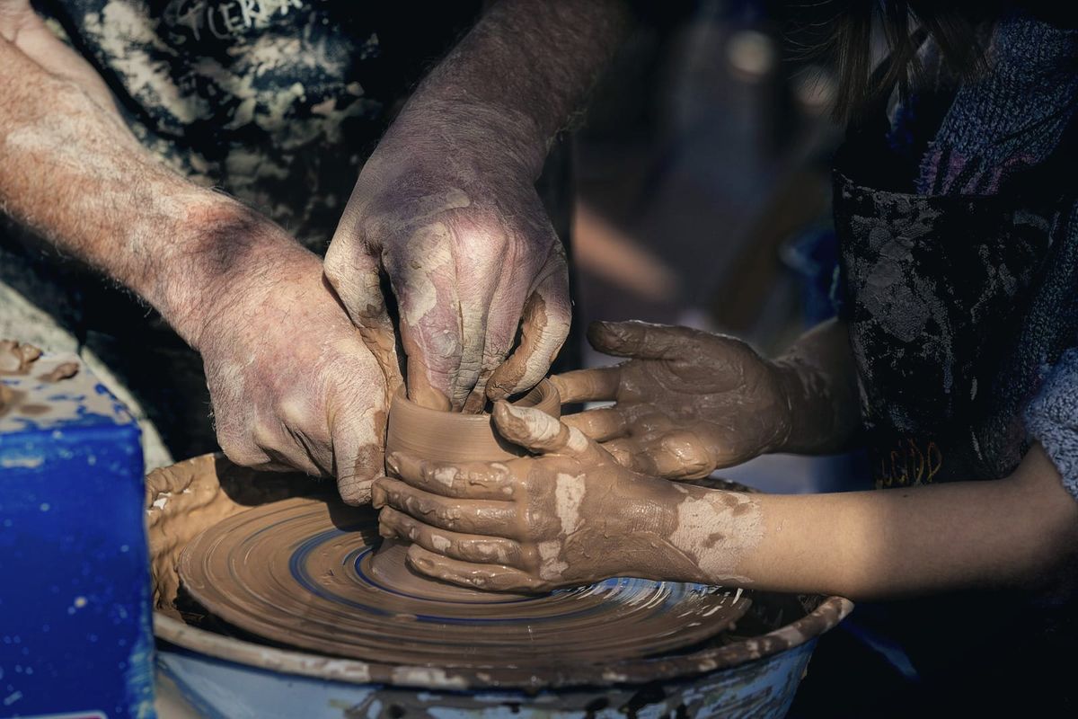 An adult potter's clay-covered hands guiding a child's hands as they shape a pot on the wheel
