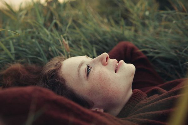 A young woman lying in long grass on a rust blanket, eyes open with a soft smile, calm and present