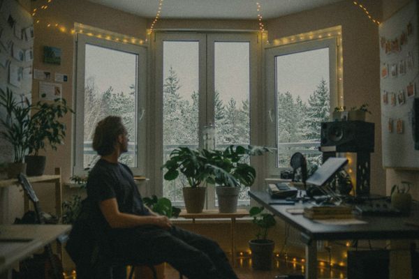 A man working at a desk in a warm, plant-filled home study with string lights, snowy trees beyond the window