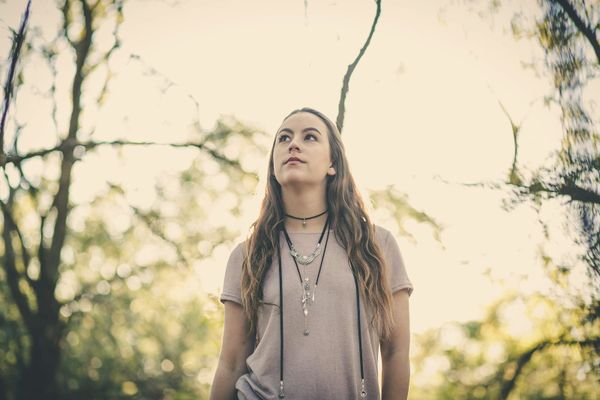 A young woman stands among trees in soft daylight, gazing up in quiet wonder.