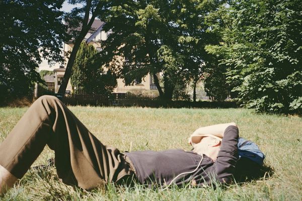 A man lying flat on a summer lawn in a park, one arm resting over his eyes