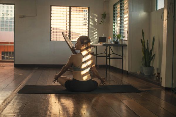 A woman sitting cross-legged on a yoga mat on a wooden floor, striped window light across her back