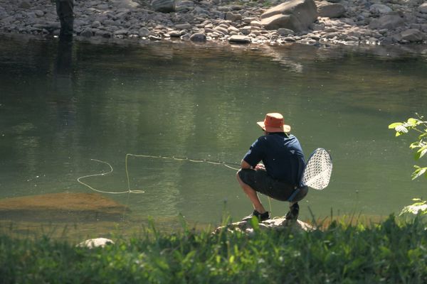 A fly fisherman crouching on a river bank, his line on the water