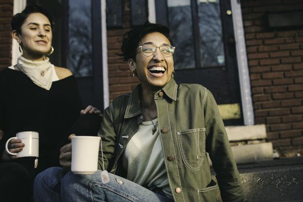 Two women laughing together on a doorstep, holding mugs of tea