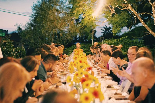 A long outdoor dinner table at dusk with people seated along it and sunflowers down the middle