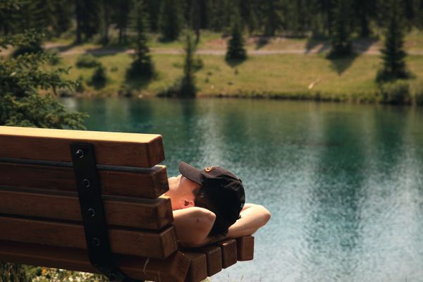 A man napping on a wooden bench beside a clear green lake