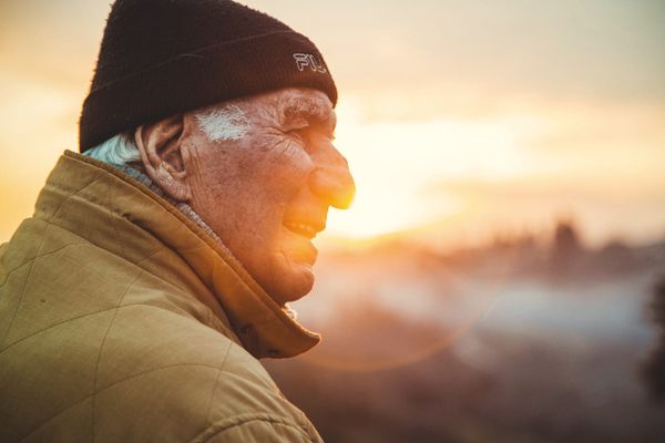An older man in a beanie smiling into the low sun over a misty city
