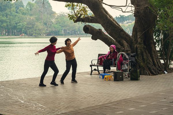 Two older women dancing slowly together by a lake while a friend watches from a bench