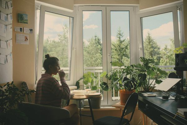 A woman drinking from a mug at a table by a sunlit window, her desk to one side