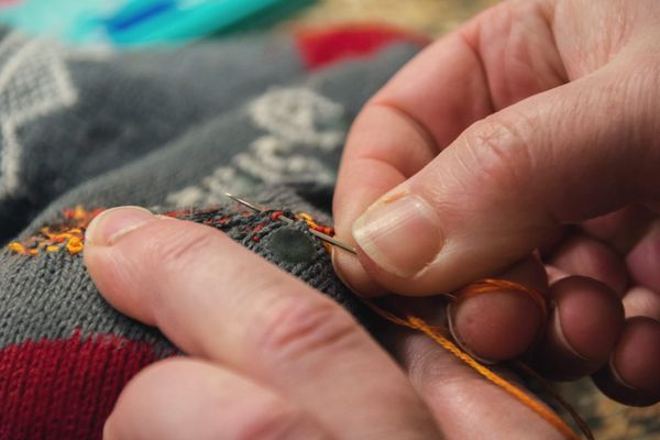 Hands darning a grey and red sock with visible orange stitching