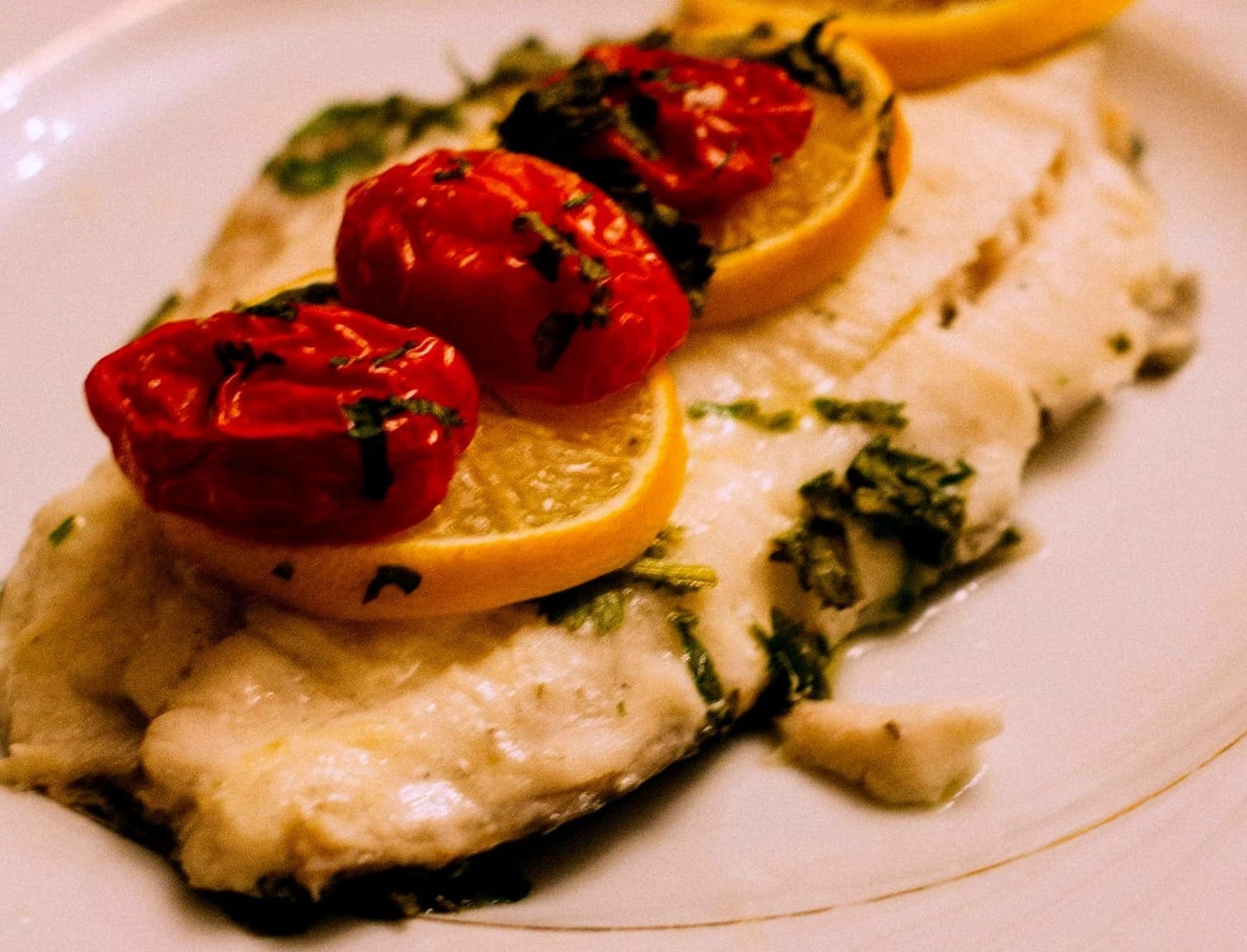 Poached fish filet topped with lemon and dried tomatoes on a white plate