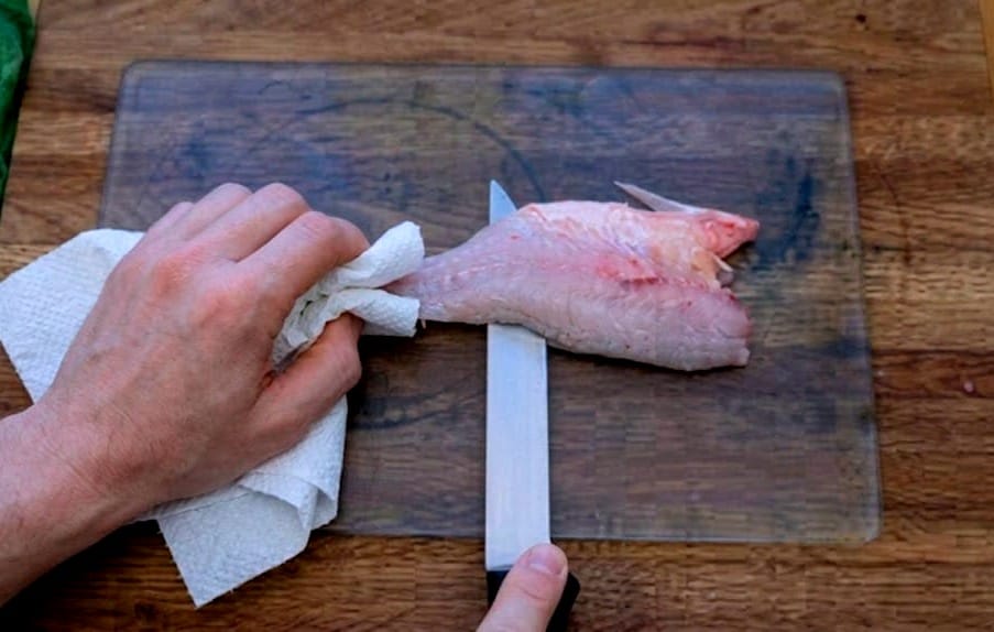 A close-up, instructional shot showing a chef's hands. One hand is holding the tail-end of a fish skin firmly with a paper towel, while the other hand holds a fillet knife angled slightly downward, separating the bright, fresh flesh from the skin.