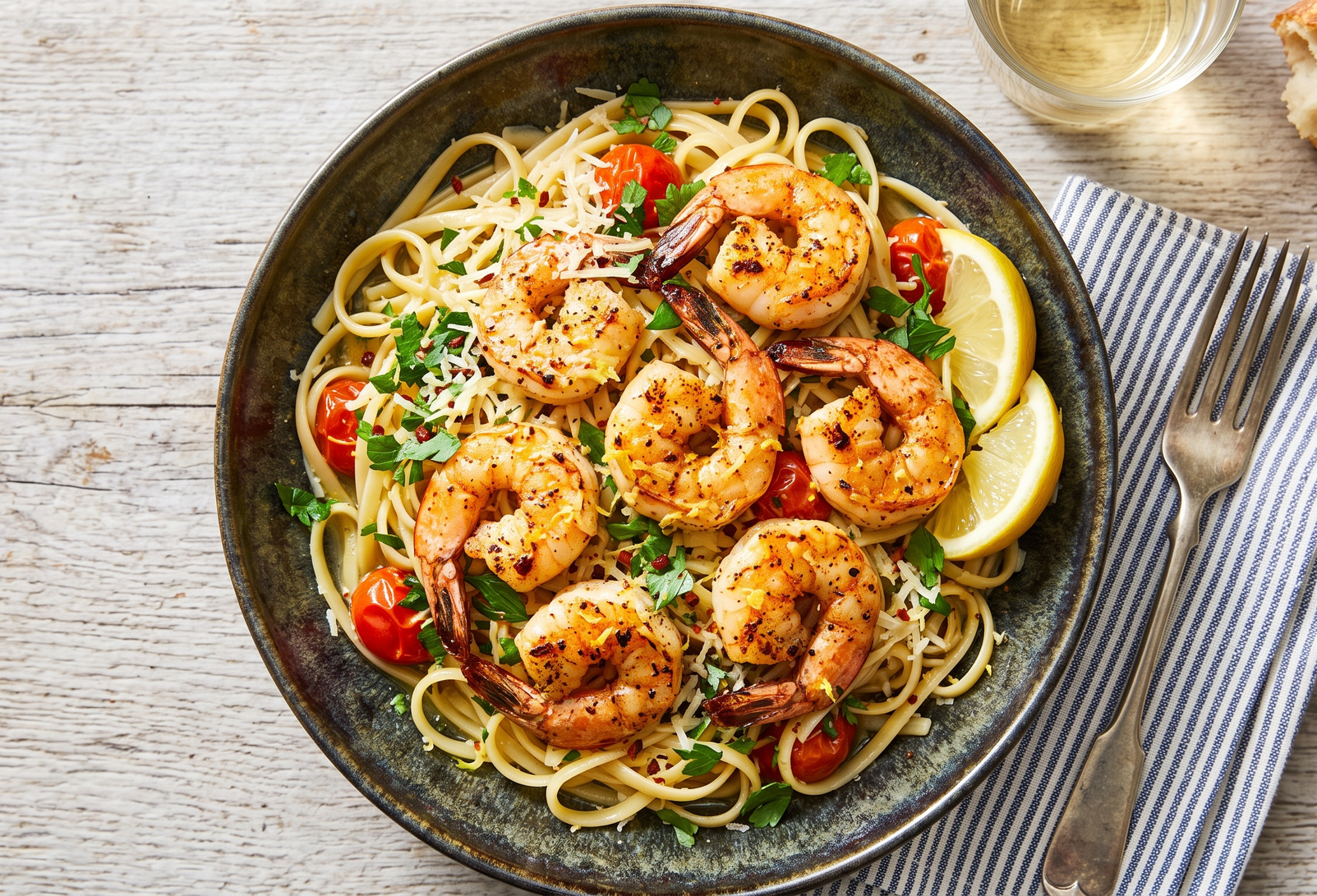 A vibrant, overhead shot of broiled shrimp over pasta in a dark ceramic dish with heirloom tomatoes, lemon wedges, and shredded parmesan cheese and fresh minced cilantro sprinkled on top.