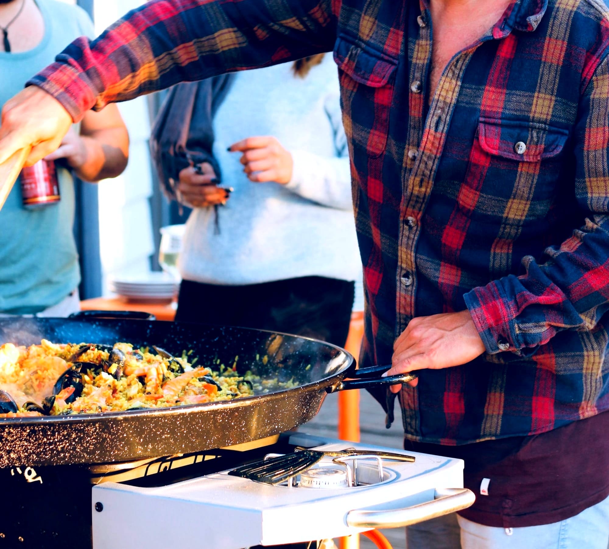 Man in a flannel shirt preparing seafood paella in a large pan with yellow rice, shrimp, and mussels on an outdoor stove top at a get together for friends who are waiting in the background.