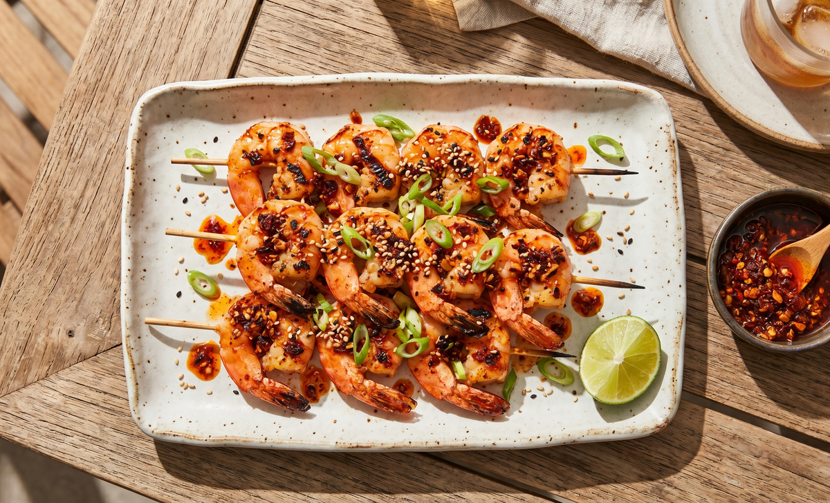 A rustic platter of grilled shrimp skewers coated in red chili crisp oil, garnished with sliced scallions and sesame seeds on an outdoor patio table.