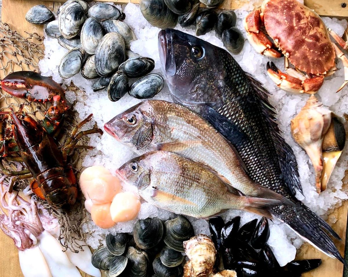 Overhead view of a wooden crate with an assortment of fresh raw seafood on top, including whole black sea bass, porgy, lobster, crab, clams, mussels, oysters, and sea scallops, over ice.