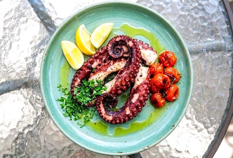 Charred octopus tentacles drizzled with olive oil and garnished with fresh parsley and lemon wedges on a rustic ceramic plate.