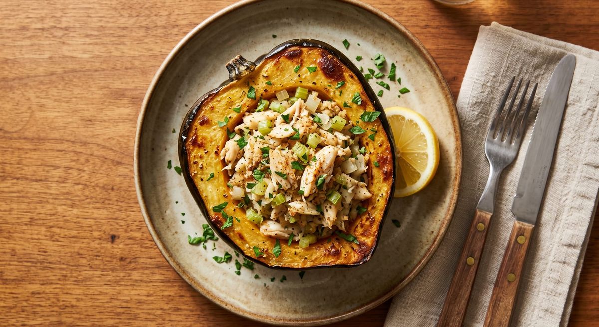 A roasted acorn squash half filled with a vibrant red and orange fresh crab meat stuffing, garnished with chopped parsley and a lemon wedge on a rustic plate.