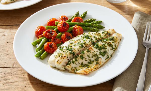 A perfectly broiled tilapia fillet topped with fresh green herbs and lemon, served on a white plate with roasted cherry tomatoes and steamed asparagus.