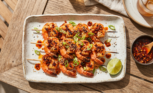 A rustic platter of grilled shrimp skewers coated in red chili crisp oil, garnished with sliced scallions and sesame seeds on an outdoor patio table.