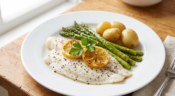 A freshly baked white flounder fillet seasoned with black pepper and topped with lemon slices and parsley, served on a white plate with roasted asparagus and baby potatoes.
