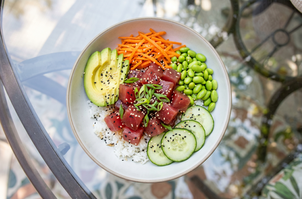 A freshly assembled poke bowl featuring deep ruby red cubes of raw Ahi tuna, bright sliced avocado, edamame, cucumber, and carrots served over warm white rice in a shallow ceramic dish.
