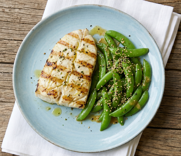 Thick, flaky grilled white halibut fillet with golden grill marks served beside bright green sugar snap peas in a miso-mustard glaze on a light blue plate.