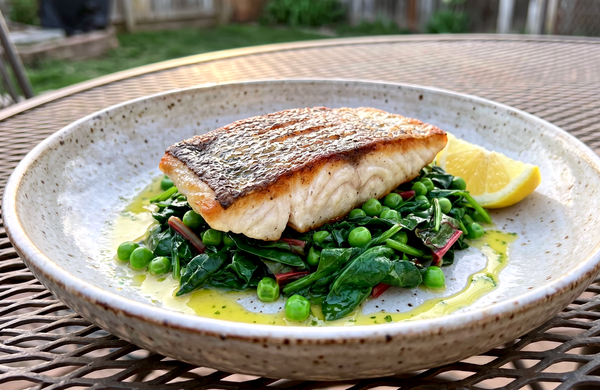 A crispy-skinned pan-seared striped bass fillet resting on a bed of bright green wilted spring greens and peas in a shallow bowl with a wedge of lemon.