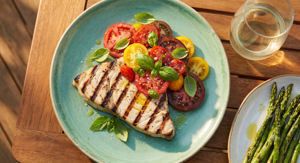 A wood-grilled swordfish steak topped with a colorful heirloom tomato and basil salad on a gree plate set on an outdoor wooden table.