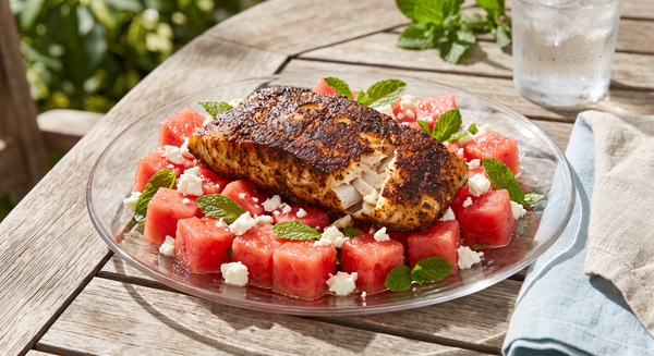 A seared blackened grouper fillet placed on top of a fresh salad made with cubed red watermelon, white feta cheese, and mint leaves on a glas plate.