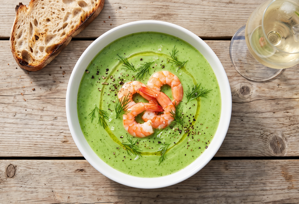 A white bowl of bright green chilled cucumber soup topped with vibrant pink poached shrimp, fresh dill, and a drizzle of olive oil on a wooden table.