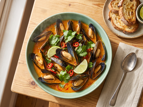 A ceramic bowl filled with steamed mussels in a vibrant orange coconut red curry broth, garnished with fresh cilantro and lime wedges on a wooden table.