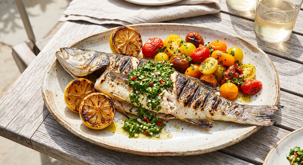 A beautifully grilled whole branzino topped with vibrant green chermoula sauce, served on a rustic platter alongside charred lemon halves and roasted heirloom tomatoes.