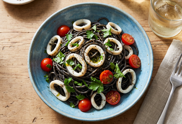 A blue ceramic bowl filled with black squid ink pasta, tossed with opaque calamari rings, red cherry tomatoes, and fresh green parsley.