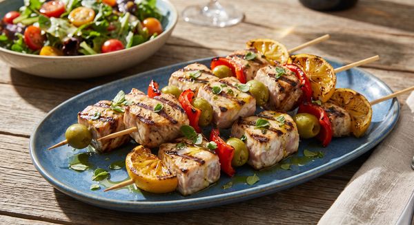 Charred swordfish skewers layered with folded lemon slices and green olives served on a rustic ceramic platter upon a wooden table.