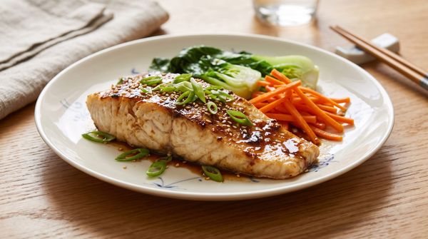 Plated roasted sea bass fillet topped with a ginger-citrus soy glaze, scallions, and sesame seeds, served alongside steamed bok choy and carrots.
