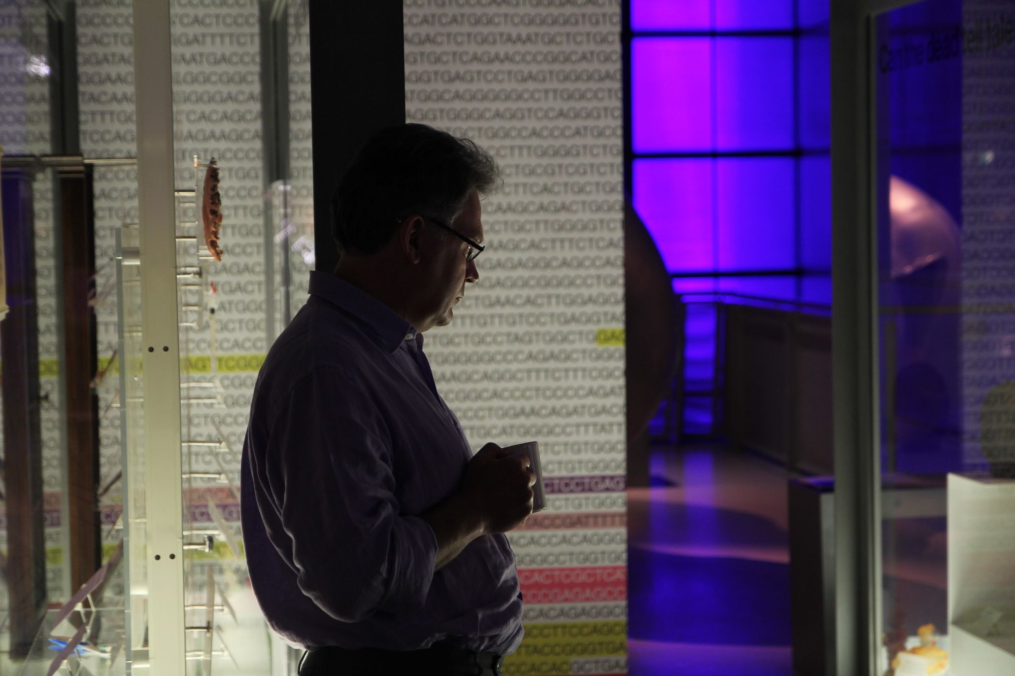 Male person holding a mug looks at a glass exhibition case, with the letters of a DNA sequence in the backdrop
