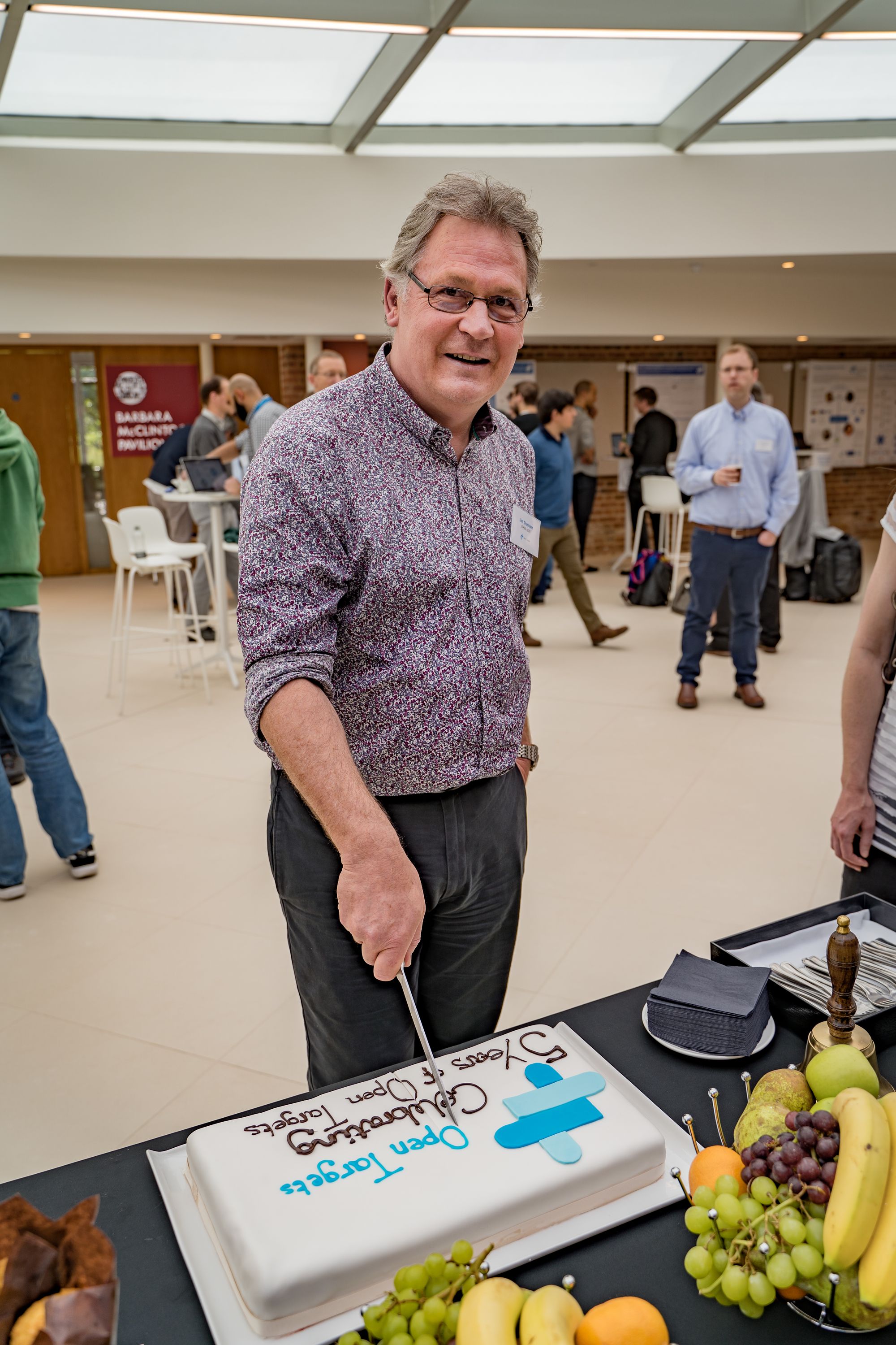 Male person stands behind a table looking at the camera, holding a knife to cut a cake.