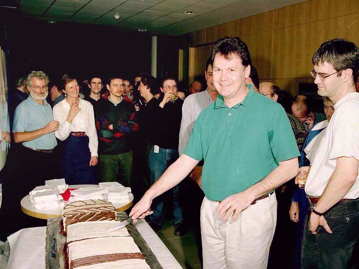 Male person, in a green shirt, stands behind a table smiling at the camera, holding a knife to cut a cake. 