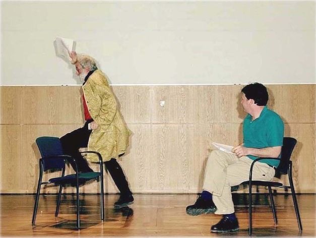 One person stepping onto a chair holding a piece of paper, another person sitting on a chair looking at the first person