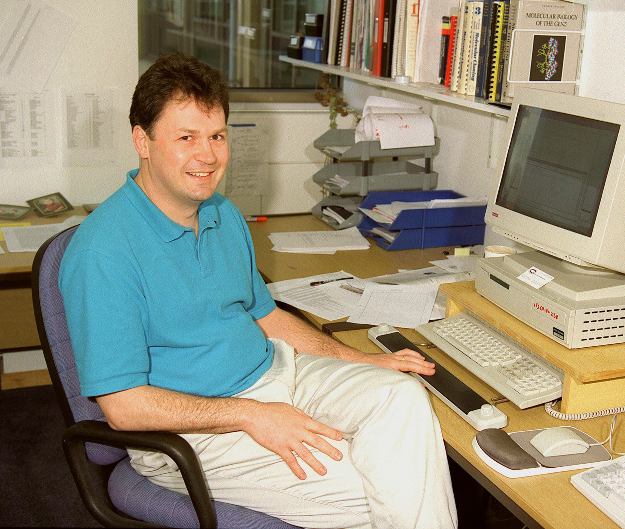 Male person in a blue polo sitting at a desk with a computer and papers, smiling at the camera