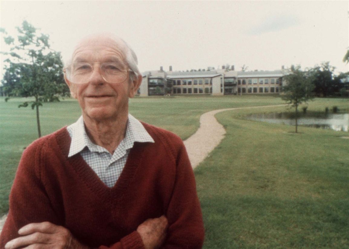 A older man in a red jumper and glasses, arms crossed and standing to the left of the image, looks into the camera. Behind him, a path leads to a long building in a park with trees and a lake.