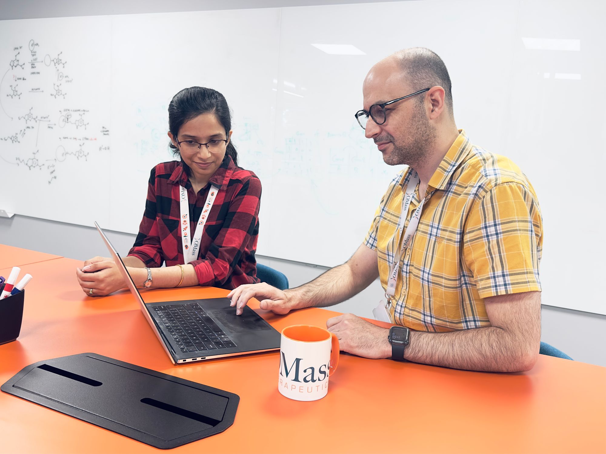 A woman and a man are seated at an orange table, looking intently at a computer which the man is manipulating. There is an OMass Therapeutics mug in front of them, and a whiteboard with chemical compounds drawn out in the background.