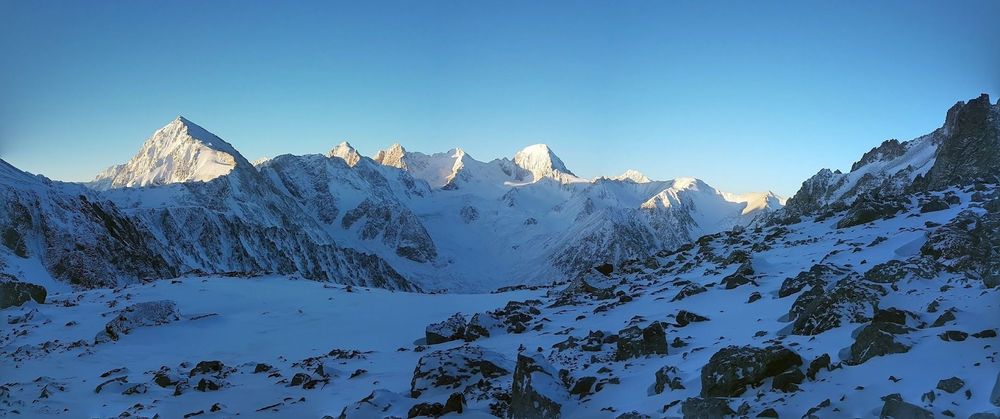 A snow-covered mountain range covered in snow. The tips of the peaks are illuminated by the sun, under a blue sky