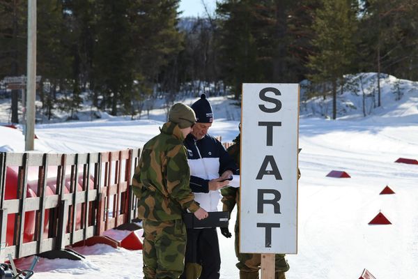 Ved et høyt START-skilt i en solfylt skiarena går en eldre mann i hvit og mørkeblå jakke gjennom papirer sammen med en soldat i feltuniform. Nypreparerte skispor og røde løypemarkeringer.