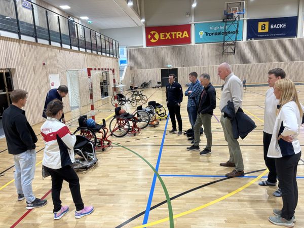 Styret i Foreningen Ridderrennet står i ring i en idrettshall. Foran dem står basketrullestoler, en racerullestol og en kjelke.